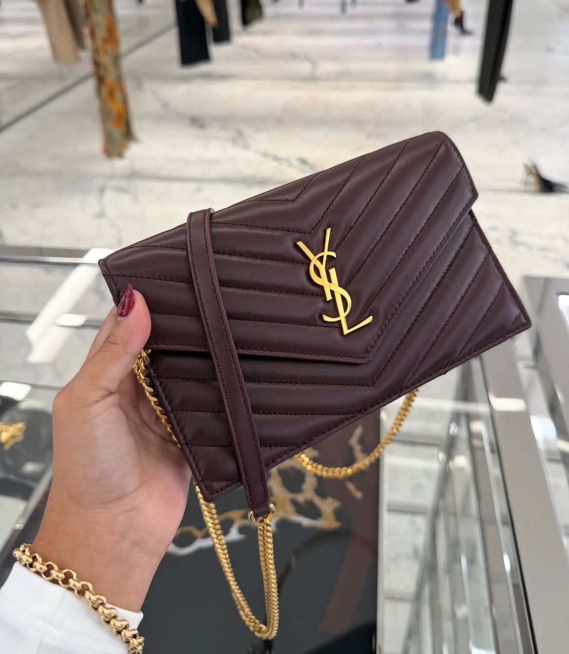 Hand holding a quilted dark brown Yves Saint Laurent wallet with a gold logo and chain strap, displayed inside a luxury store.