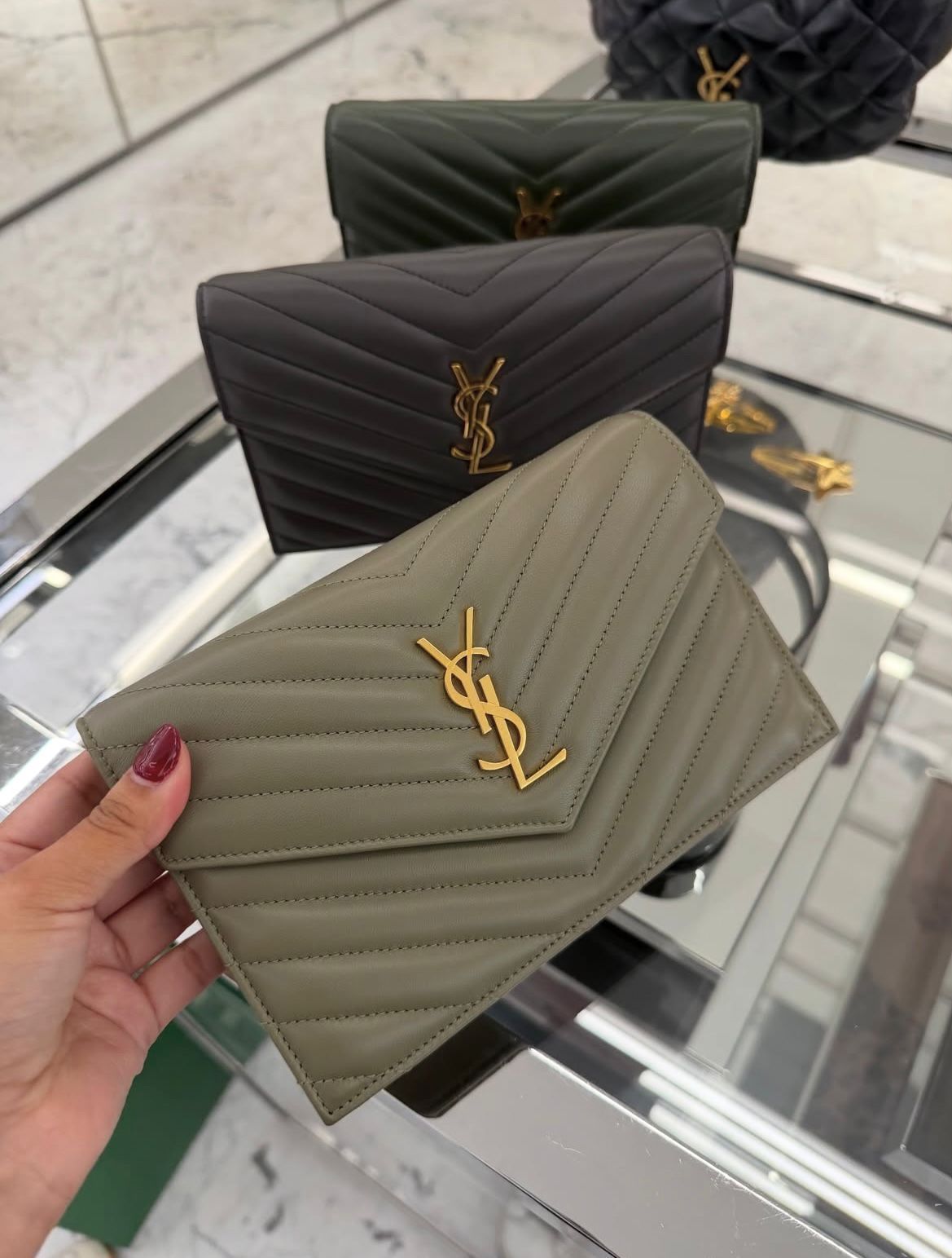 Hand holding a quilted khaki Yves Saint Laurent wallet with a gold logo, with black and dark green wallets displayed on a glass shelf in the background.
