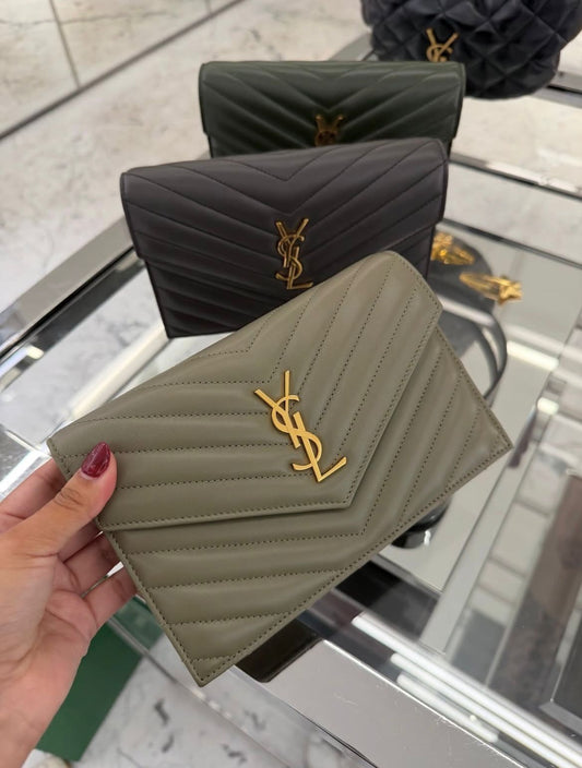 Hand holding a quilted khaki Yves Saint Laurent wallet with a gold logo, with black and dark green wallets displayed on a glass shelf in the background.